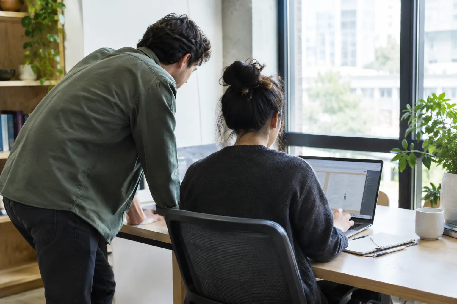 Une personne observe une collègue au travail, un carnet ouvert près de son poste.